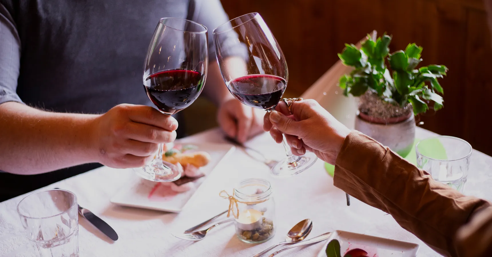 Paar stößt mit Rotwein an, weiß gedeckter Tisch mit Kerzenlicht in holzvertäfelter Stube