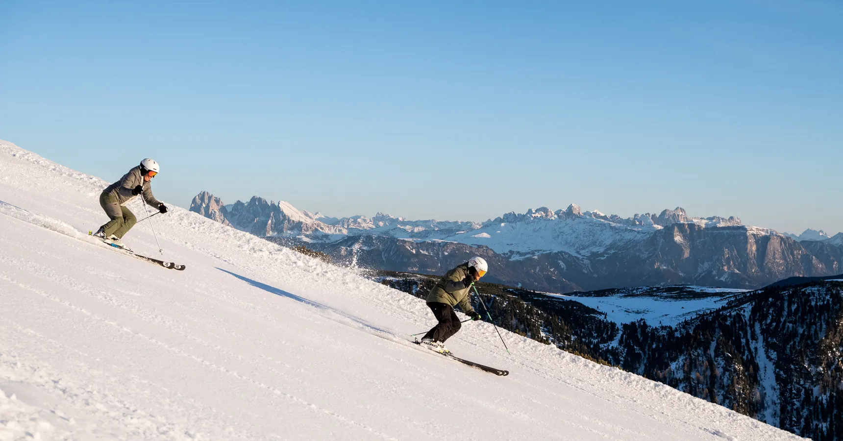 Zwei Skifahrer auf präparierter Piste mit Blick auf die Dolomiten bei blauem Himmel