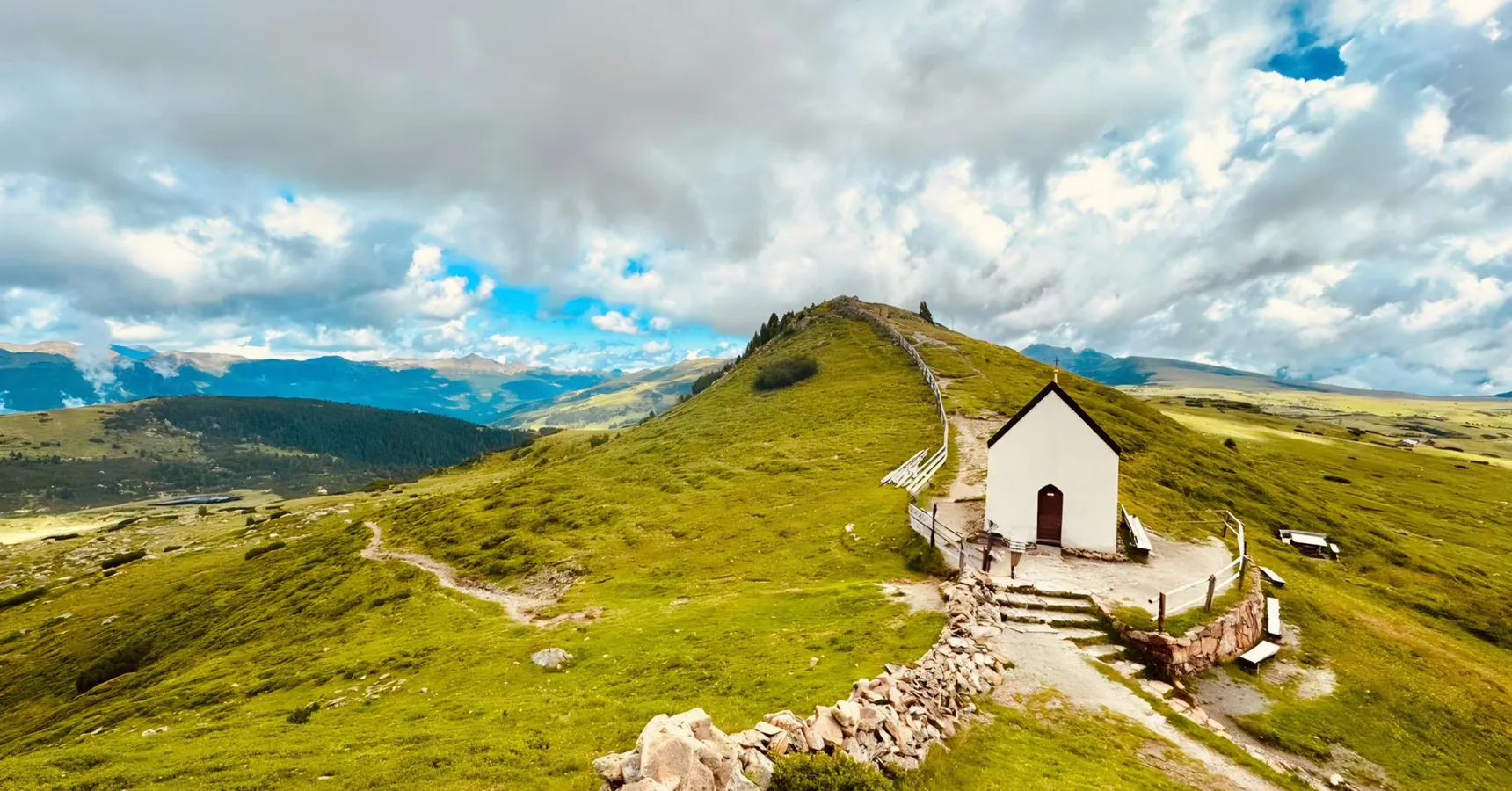 Kleine weiße Bergkapelle auf grünem Höhenrücken mit Wanderweg und Wolkenhimmel im Sarntal