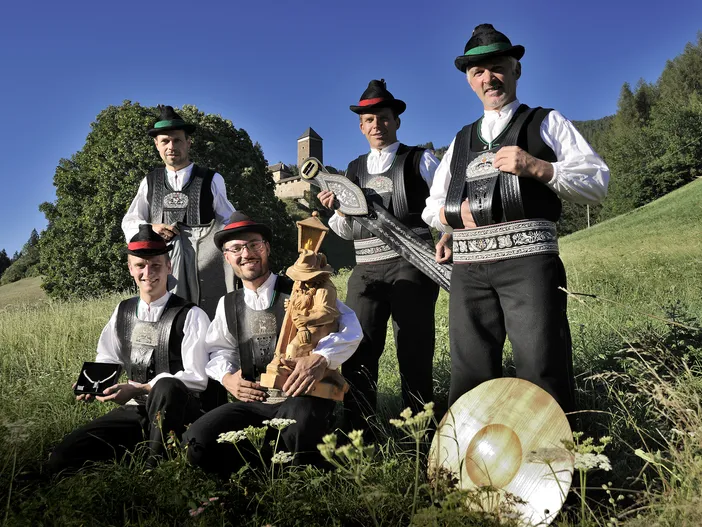 Männer in traditioneller Sarner Tracht mit Hut und Schnitzarbeiten auf sommerlicher Bergwiese