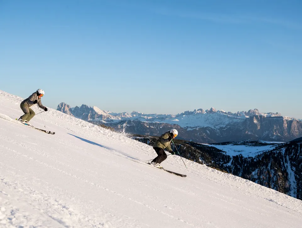 Zwei Skifahrer auf präparierter Piste mit Blick auf die Dolomiten bei blauem Himmel