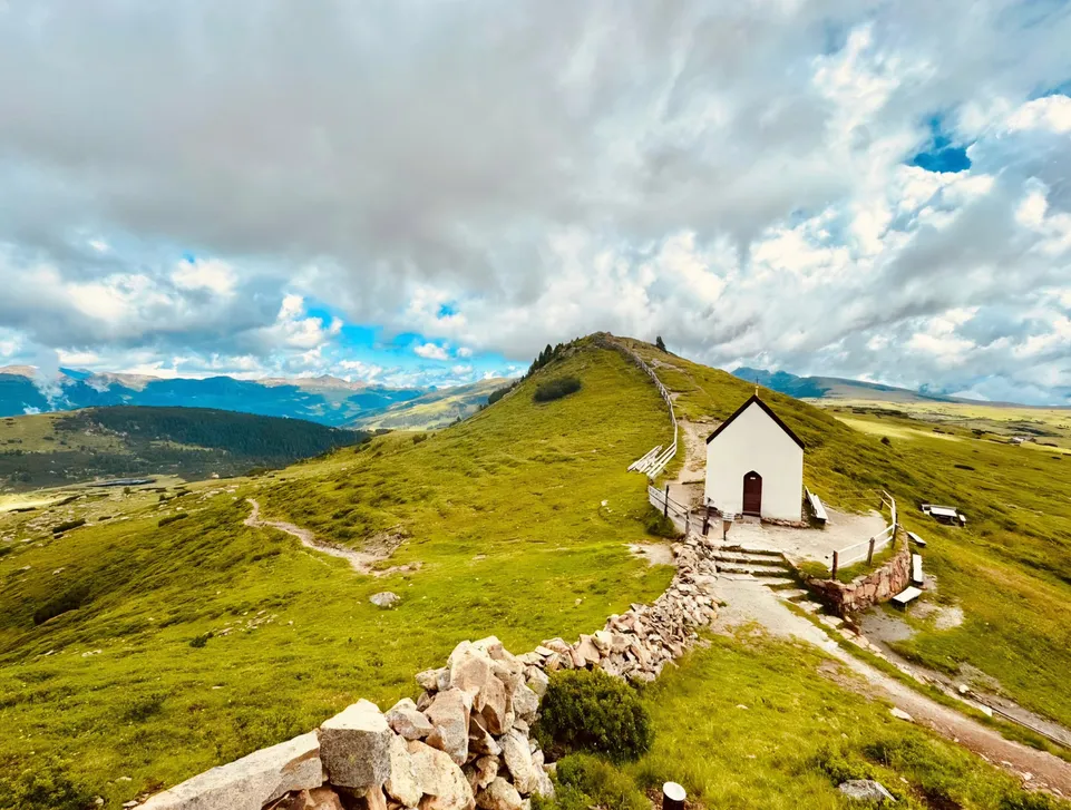 Kleine weiße Bergkapelle auf grünem Höhenrücken mit Wanderweg und Wolkenhimmel im Sarntal