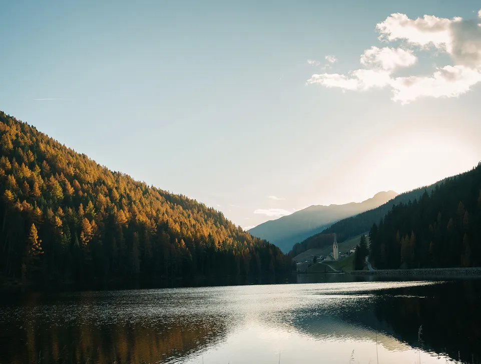 Herbstlicher Bergsee im Sarntal mit goldgelben Lärchen und Kirchturm bei Sonnenuntergang
