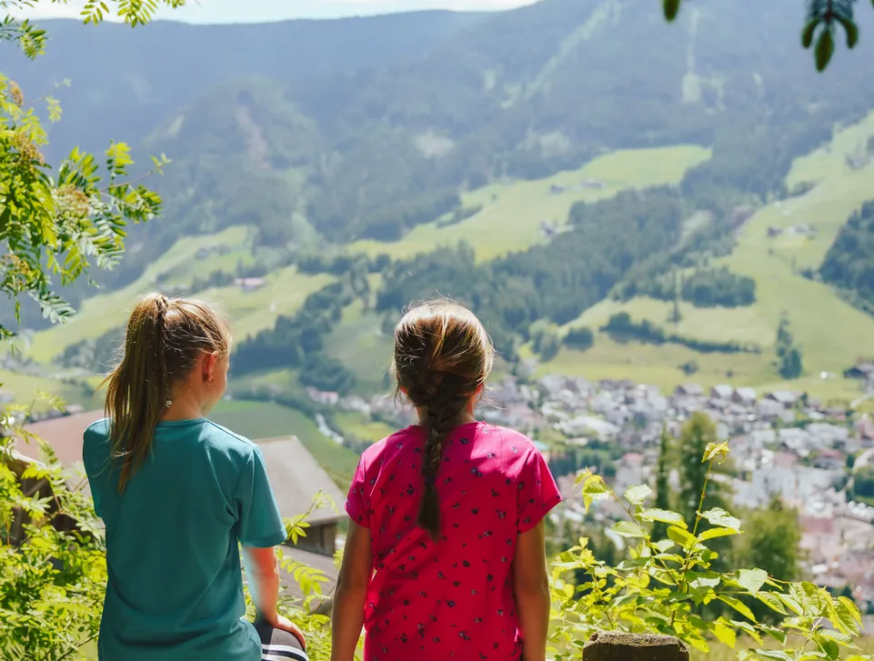 Zwei Mädchen sitzen auf einem Holzzaun und blicken auf das grüne Dorf Sarnthein im Tal